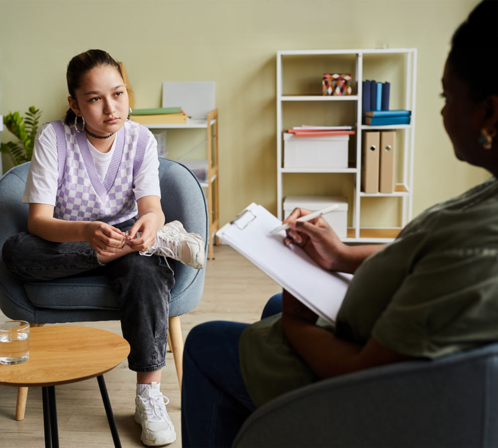 Teenage girl sitting on armchair with clinician taking notes on paper during session at office